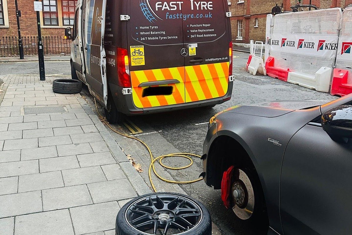 A dark grey car at the kerb with its front wheel off and the brake disc exposed, the removed wheel on the pavement and an air line running back to the Fast Tyre van